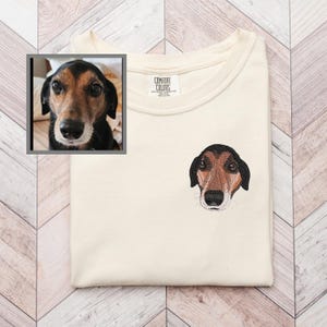 May include: Cream-colored t-shirt with an embroidered dog face on the chest. The dog has brown and black fur with a black nose and is facing forward. A framed photo of the dog is in the upper left corner. The shirt has a Comfort Colors tag.