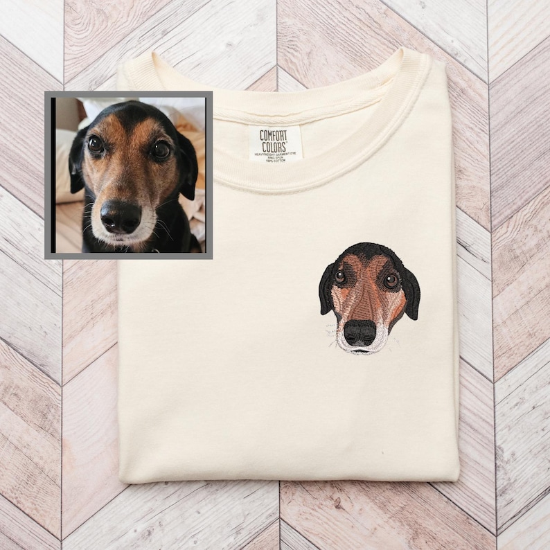 May include: Cream-colored t-shirt with an embroidered dog face on the chest. The dog has brown and black fur with a black nose and is facing forward. A framed photo of the dog is in the upper left corner. The shirt has a Comfort Colors tag.