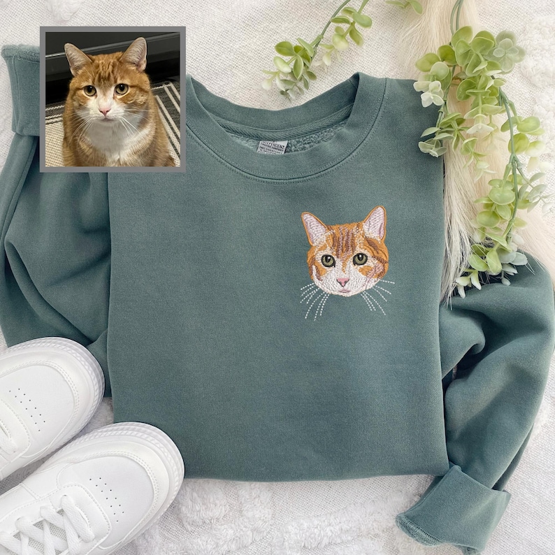 Puede incluir: Una sudadera verde con una imagen bordada de la cara de un gato en la parte delantera. El gato tiene pelo naranja y blanco.