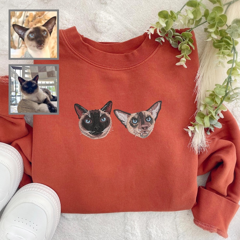 Puede incluir: Una sudadera de color &oacute;xido con dos im&aacute;genes bordadas de gatos siameses en la parte delantera. Los gatos est&aacute;n mirando hacia adelante con los ojos abiertos.