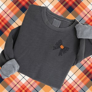 Puede incluir: Una sudadera gris oscuro con un lazo de encaje negro con una calabaza naranja en el centro. La sudadera est&aacute; colocada sobre un fondo a cuadros.