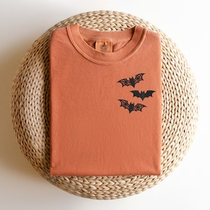 May include: A folded orange t-shirt with a black bat design on the front. The t-shirt is on a woven straw surface. The text "COMFORT COLORS" is visible on the tag.