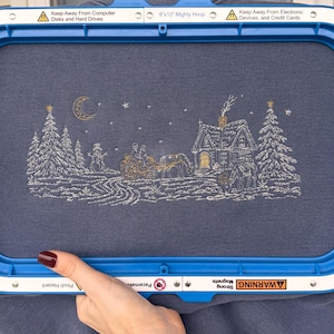 May include: A blue fabric held in an 8x13 inch Mighty Hoop frame featuring an embroidered winter scene with a house, trees, a snowman, and a horse-drawn sleigh under a crescent moon. The text on the frame reads Keep Away From Computer Disks and Hard Drives, 8x13 Mighty Hoop, Keep Away From Electronic Devices, and Credit Cards.