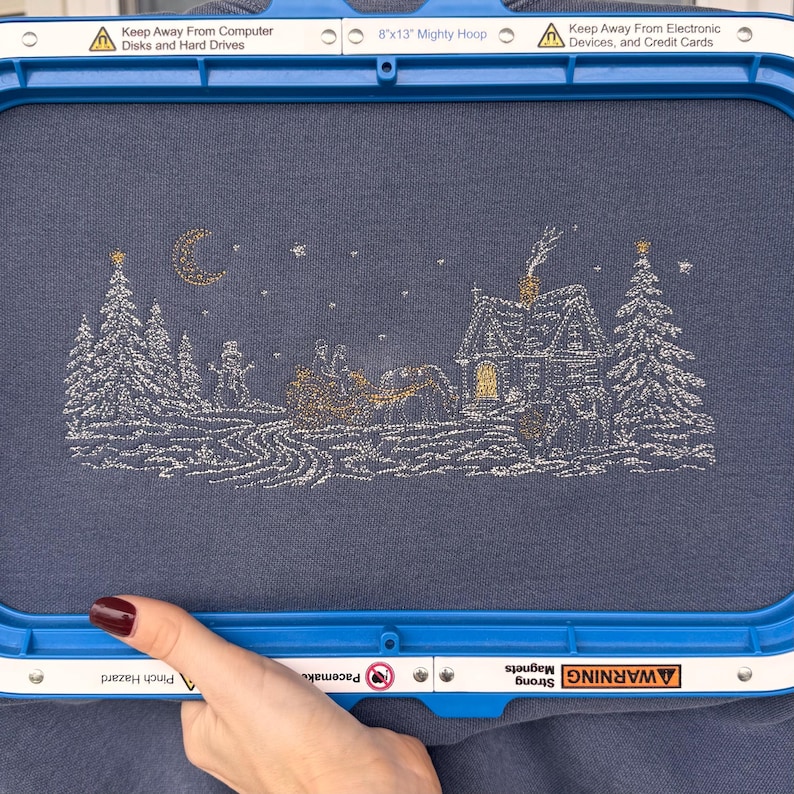 May include: A blue fabric held in an 8x13 inch Mighty Hoop frame featuring an embroidered winter scene with a house, trees, a snowman, and a horse-drawn sleigh under a crescent moon. The text on the frame reads Keep Away From Computer Disks and Hard Drives, 8x13 Mighty Hoop, Keep Away From Electronic Devices, and Credit Cards.