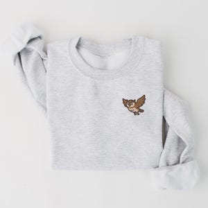 以下が含まれることがあります： 胸に小さなフクロウの刺繍パッチが付いた、明るい灰色のスウェットシャツ。フクロウは茶色とタン色で、翼を広げています。袖口は折り返されており、スウェットシャツは折りたたまれています。