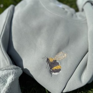 May include: A light green sweatshirt with an embroidered bumble bee design. The bee is yellow and black with brown wings.