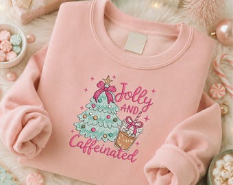 Puede incluir: Una sudadera de cuello redondo rosa claro con un estampado gráfico de un árbol de Navidad estilizado con un lazo rosa y una taza de café con un lazo rosa y una pajita. El texto dice Jolly AND Caffeinated en letra cursiva rosa con estrellas.