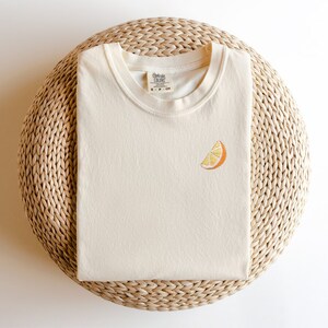 Puede incluir: Una camiseta de color crema con una peque&ntilde;a rodaja de naranja bordada en la parte delantera. La camiseta est&aacute; sobre una estera de paja tejida.