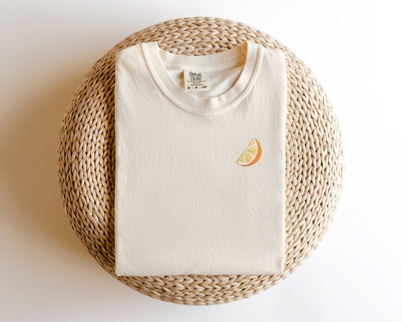 Puede incluir: Una camiseta de color crema con una peque&ntilde;a rodaja de naranja bordada en la parte delantera. La camiseta est&aacute; sobre una estera de paja tejida.