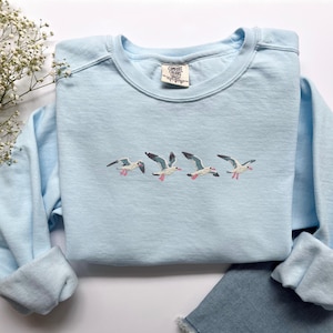 Puede incluir: Sudadera azul claro con gaviotas bordadas en vuelo. Las aves tienen cuerpos blancos, alas grises y patas rosas. La sudadera tiene cuello redondo y mangas largas. Una peque&ntilde;a etiqueta es visible en el escote.