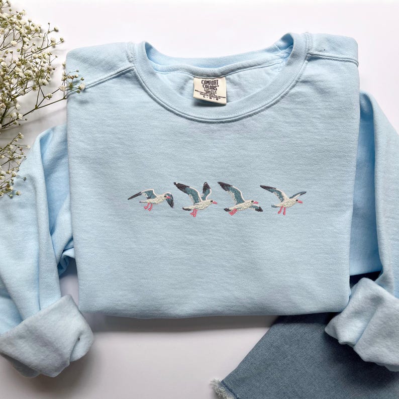 Puede incluir: Sudadera azul claro con gaviotas bordadas en vuelo. Las aves tienen cuerpos blancos, alas grises y patas rosas. La sudadera tiene cuello redondo y mangas largas. Una peque&ntilde;a etiqueta es visible en el escote.