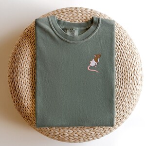 May include: A green t-shirt with a small embroidered rat design. The t-shirt has a crew neck and is made of a soft, comfortable fabric. The text "COMFORT COLORS" is visible on the tag.