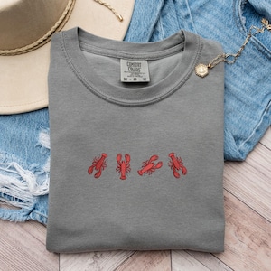 May include: A gray t-shirt with a row of five red embroidered lobsters. The shirt has a crew neck and a tag that reads "Comfort Colors".