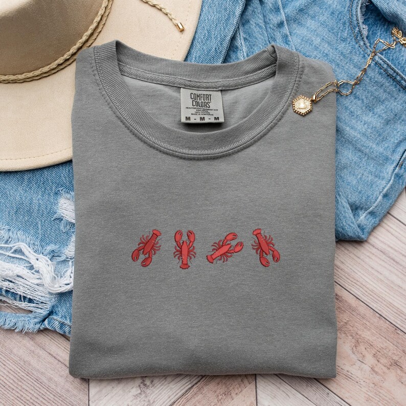 May include: A gray t-shirt with a row of five red embroidered lobsters. The shirt has a crew neck and a tag that reads "Comfort Colors".