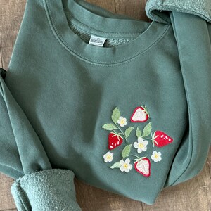May include: A green sweatshirt with an embroidered design of strawberries and white flowers. The sweatshirt has a crew neck and long sleeves.