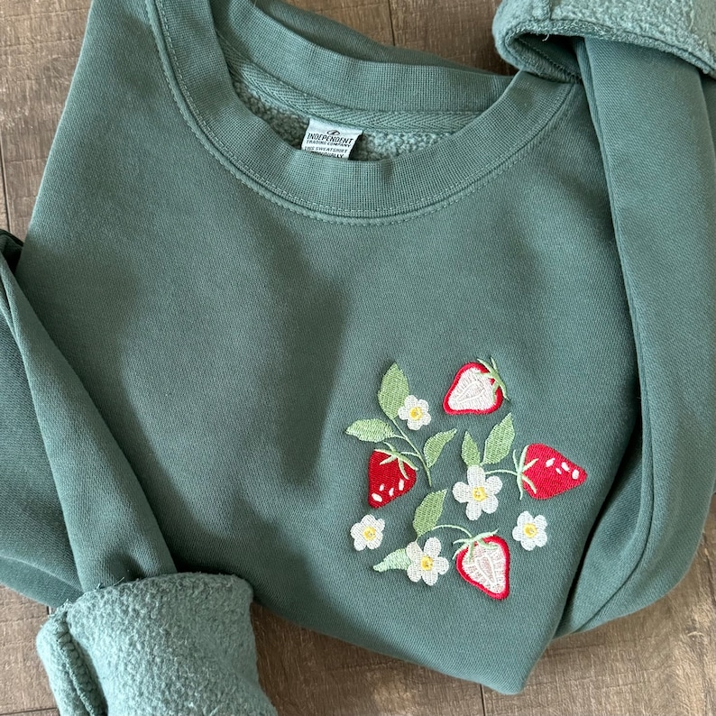May include: A green sweatshirt with an embroidered design of strawberries and white flowers. The sweatshirt has a crew neck and long sleeves.