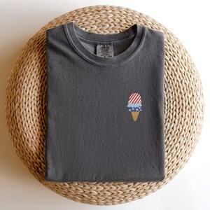 May include: A dark gray t-shirt with a small embroidered ice cream cone design. The ice cream cone features red and white stripes, blue stars, and a tan cone. The shirt is folded neatly on a woven, circular surface.