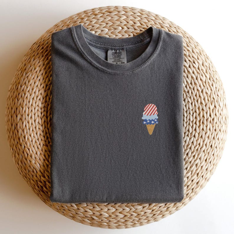 May include: A dark gray t-shirt with a small embroidered ice cream cone design. The ice cream cone features red and white stripes, blue stars, and a tan cone. The shirt is folded neatly on a woven, circular surface.