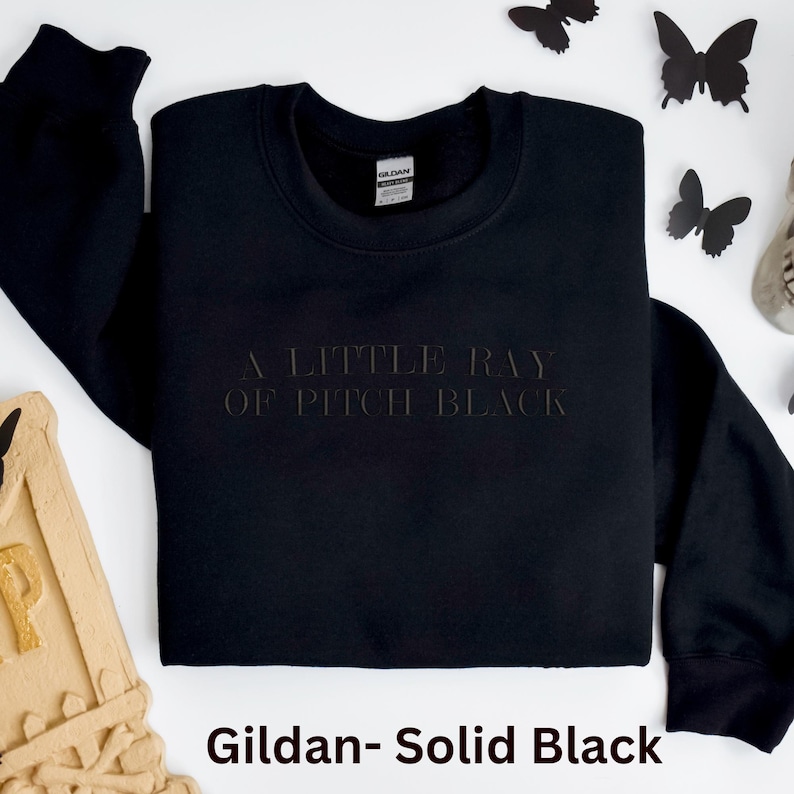 May include: A black sweatshirt with the phrase "A Little Ray of Pitch Black" embossed on the front. The sweatshirt is a solid black color and is made by Gildan. Decorative black butterfly cutouts are scattered around the sweatshirt.