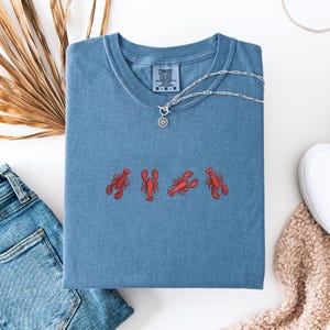 May include: A blue t-shirt with a red embroidered design of five lobsters. The shirt has a crew neck and short sleeves. The brand label says "Comfort Colors".