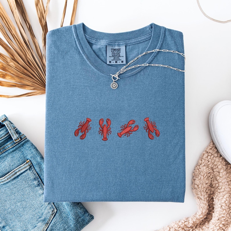 May include: A blue t-shirt with a red embroidered design of five lobsters. The shirt has a crew neck and short sleeves. The brand label says "Comfort Colors".