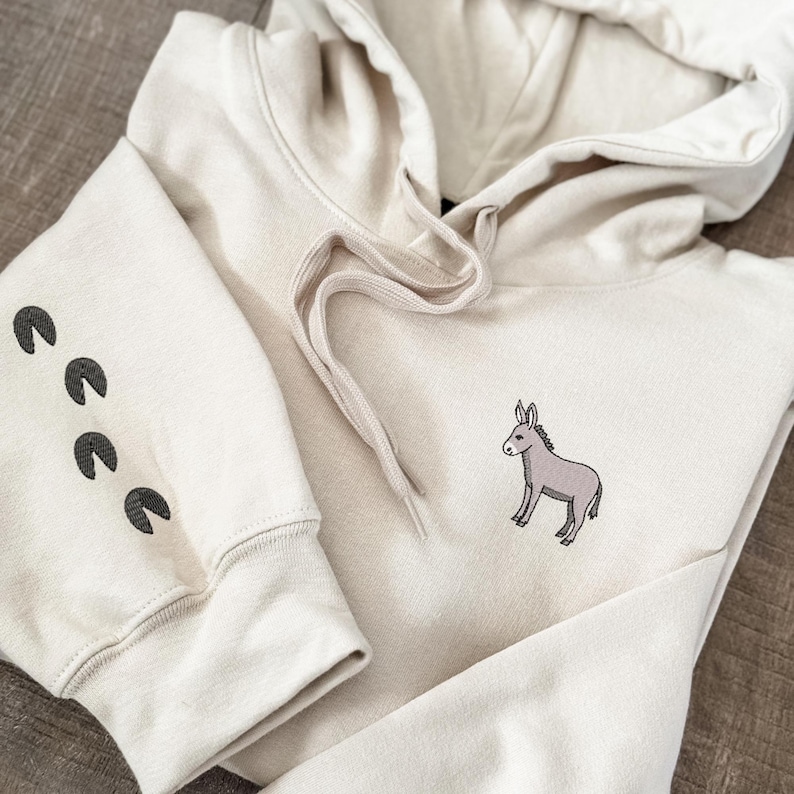 Puede incluir: Una sudadera con capucha de color crema con un peque&ntilde;o burro bordado en la parte delantera. La sudadera tiene una capucha con cord&oacute;n y un bolsillo canguro. La manga izquierda tiene cuatro huellas de patas negras.