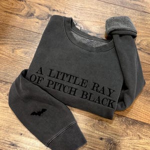 May include: Dark gray sweatshirt with the text "A LITTLE RAY OF PITCH BLACK" in black lettering. A small bat graphic is on the left sleeve. The sweatshirt has a crew neck and a fleece lining.