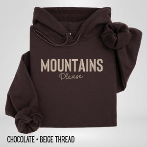 May include: A dark brown hoodie with the words "MOUNTAINS Please" in beige thread. The hoodie has a drawstring hood and long sleeves with cuffed wrists. The text "CHOCOLATE + BEIGE THREAD" is at the bottom.
