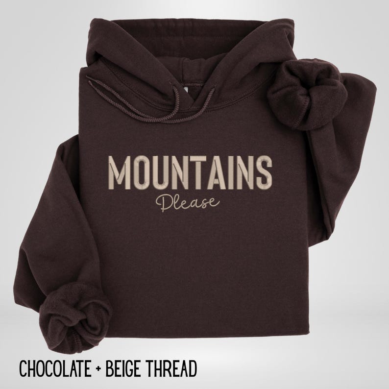 May include: A dark brown hoodie with the words "MOUNTAINS Please" in beige thread. The hoodie has a drawstring hood and long sleeves with cuffed wrists. The text "CHOCOLATE + BEIGE THREAD" is at the bottom.