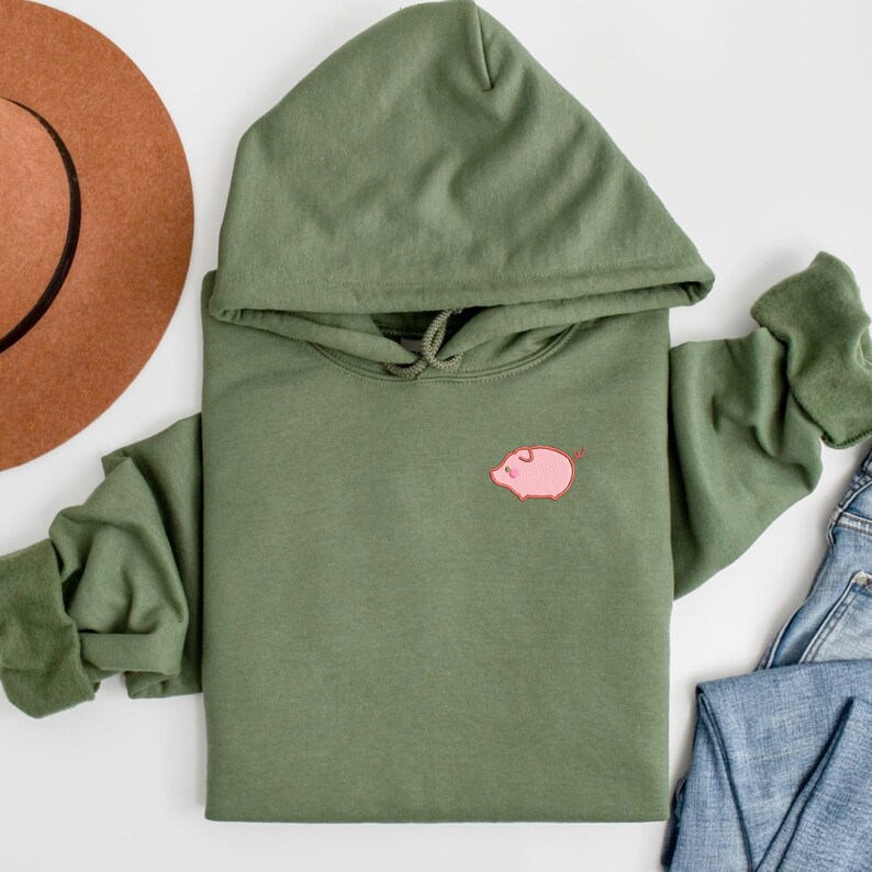 K&ouml;nnte beinhalten: Ein gr&uuml;ner Kapuzenpullover mit einem gestickten rosa Schweinchen auf der Vorderseite.