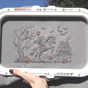 以下が含まれることがあります： 白い刺繍枠に収められたグレーの布地。黒とオレンジの糸で、馬に乗った首のない騎士、フクロウのいる木、家、墓石、コウモリ、三日月、3つのカボチャが詳細に刺繍されています。
