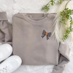 Puede incluir: Una sudadera marr&oacute;n claro con un lazo de encaje negro con una peque&ntilde;a calabaza naranja en el centro. La sudadera est&aacute; sobre una superficie blanca y peluda.