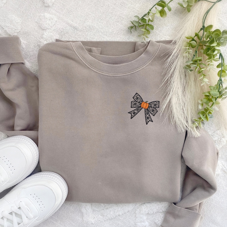 Puede incluir: Una sudadera marr&oacute;n claro con un lazo de encaje negro con una peque&ntilde;a calabaza naranja en el centro. La sudadera est&aacute; sobre una superficie blanca y peluda.