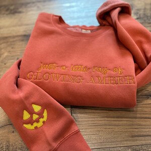 May include: A terracotta-colored sweatshirt with the embroidered text "just a little ray of GLOWING AMBER." The left sleeve features a yellow embroidered jack-o'-lantern face. The sweatshirt is folded on a wooden surface.