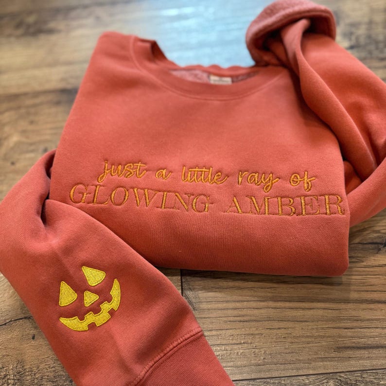 May include: A terracotta-colored sweatshirt with the embroidered text "just a little ray of GLOWING AMBER." The left sleeve features a yellow embroidered jack-o'-lantern face. The sweatshirt is folded on a wooden surface.