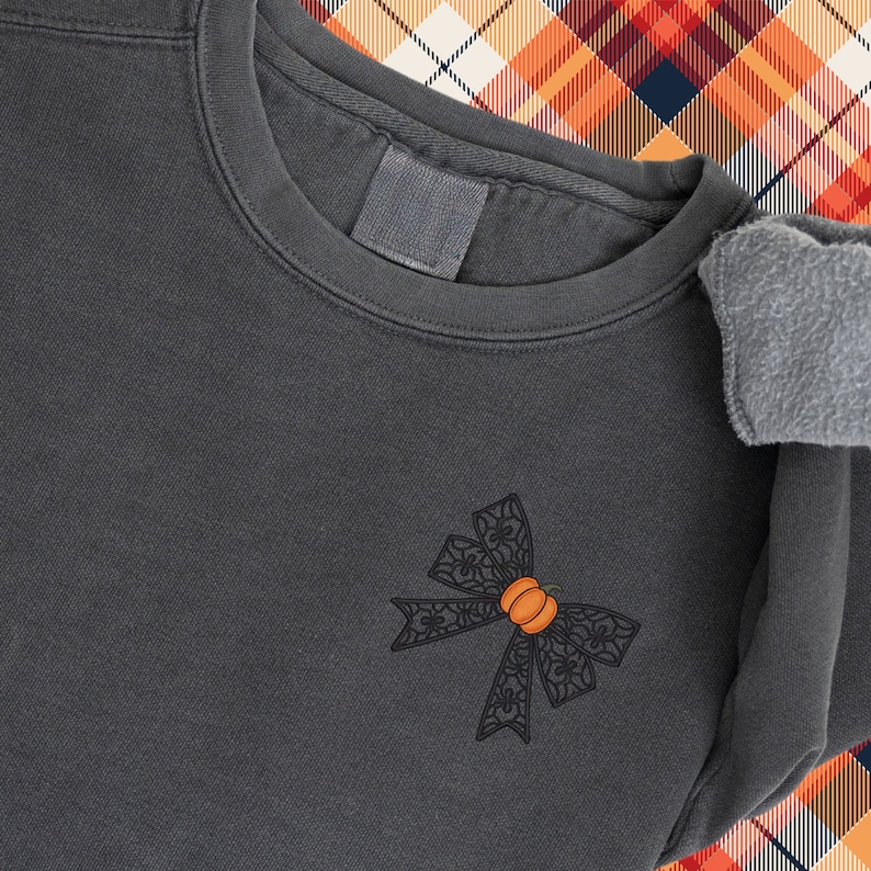 Puede incluir: Una sudadera gris oscura con un lazo de encaje negro con una peque&ntilde;a calabaza naranja en el centro. La sudadera est&aacute; sobre una tela a cuadros.