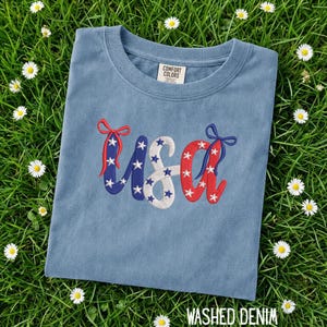 Pu&ograve; includere: Una maglietta blu con la scritta "USA" ricamata in rosso, bianco e blu con dettagli a stella. La maglietta &egrave; realizzata in materiale morbido ed &egrave; etichettata "Washed Denim". La maglietta &egrave; su erba verde con piccoli fiori bianchi.