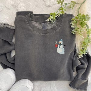 May include: A charcoal gray sweatshirt with a small embroidered snowman design on the chest. The snowman wears a blue hat and scarf and holds a red mug. The sweatshirt is folded with white sneakers visible.