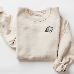 May include: A beige sweatshirt with a small embroidered possum on the left chest. The possum is gray with pink ears and a black nose.