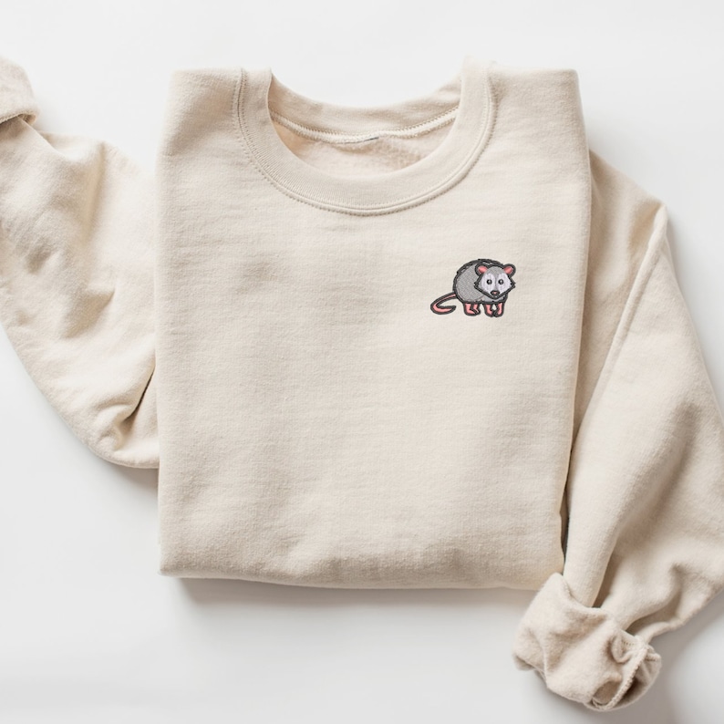 May include: A beige sweatshirt with a small embroidered possum on the left chest. The possum is gray with pink ears and a black nose.