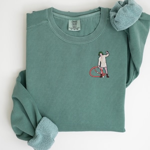 Może przedstawiać: Zielono-niebieska bluza z haftowanym białym obrazkiem osoby trzymającej czerwone k&oacute;łko. Bluza ma metkę Comfort Colors w lewym dolnym rogu.