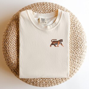 May include: A cream colored t-shirt with a small embroidered tiger design. The t-shirt is on a woven straw mat.