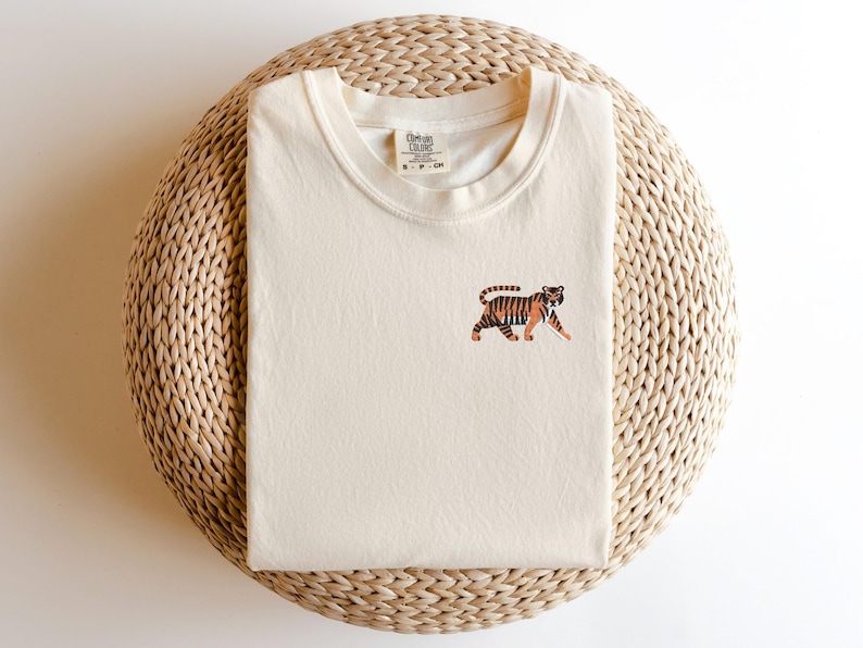 May include: A cream colored t-shirt with a small embroidered tiger design. The t-shirt is on a woven straw mat.