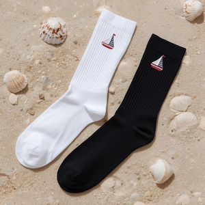 May include: A pair of crew socks, one white and one black, each embroidered with a sailboat design. The socks are displayed on a sandy surface with seashells scattered around them. The sailboat design features a red and blue striped sail.