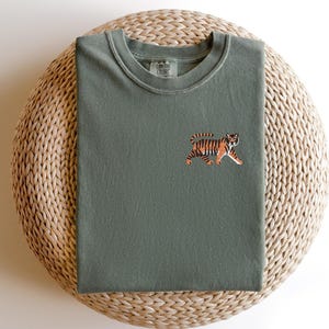 May include: A green t-shirt with an embroidered tiger design. The tiger is orange and black with a white background.