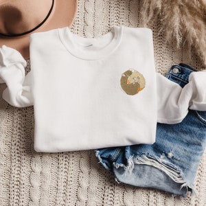 May include: White sweatshirt with a brown and beige embroidered design of two animals curled up together. The sweatshirt is laying on a beige knitted surface with a pair of blue denim shorts.