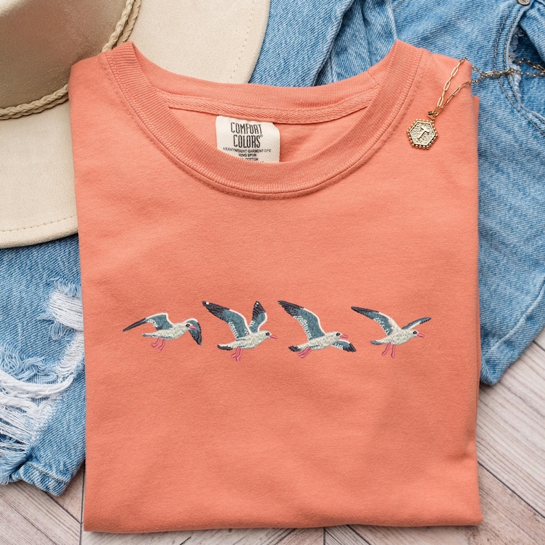 Puede incluir: Una camiseta de color coral con un dise&ntilde;o bordado de cinco gaviotas volando en fila. La camiseta tiene cuello redondo y la etiqueta de la marca "COMFORT COLORS" es visible en la parte delantera.