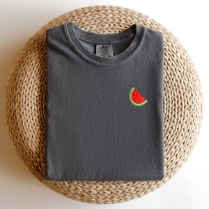 Puede incluir: Una camiseta gris oscuro con una peque&ntilde;a rodaja de sand&iacute;a bordada en la parte delantera.
