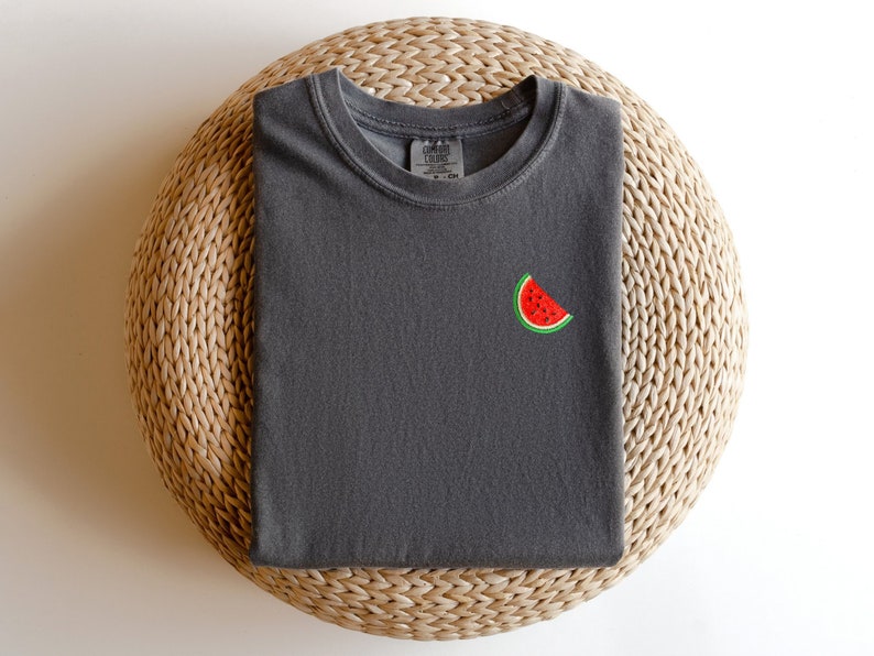 Puede incluir: Una camiseta gris oscuro con una peque&ntilde;a rodaja de sand&iacute;a bordada en la parte delantera.