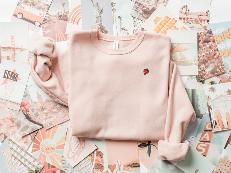 May include: A pale pink sweatshirt with a small embroidered strawberry on the front. The sweatshirt is laid out on a background of colorful patterned paper.